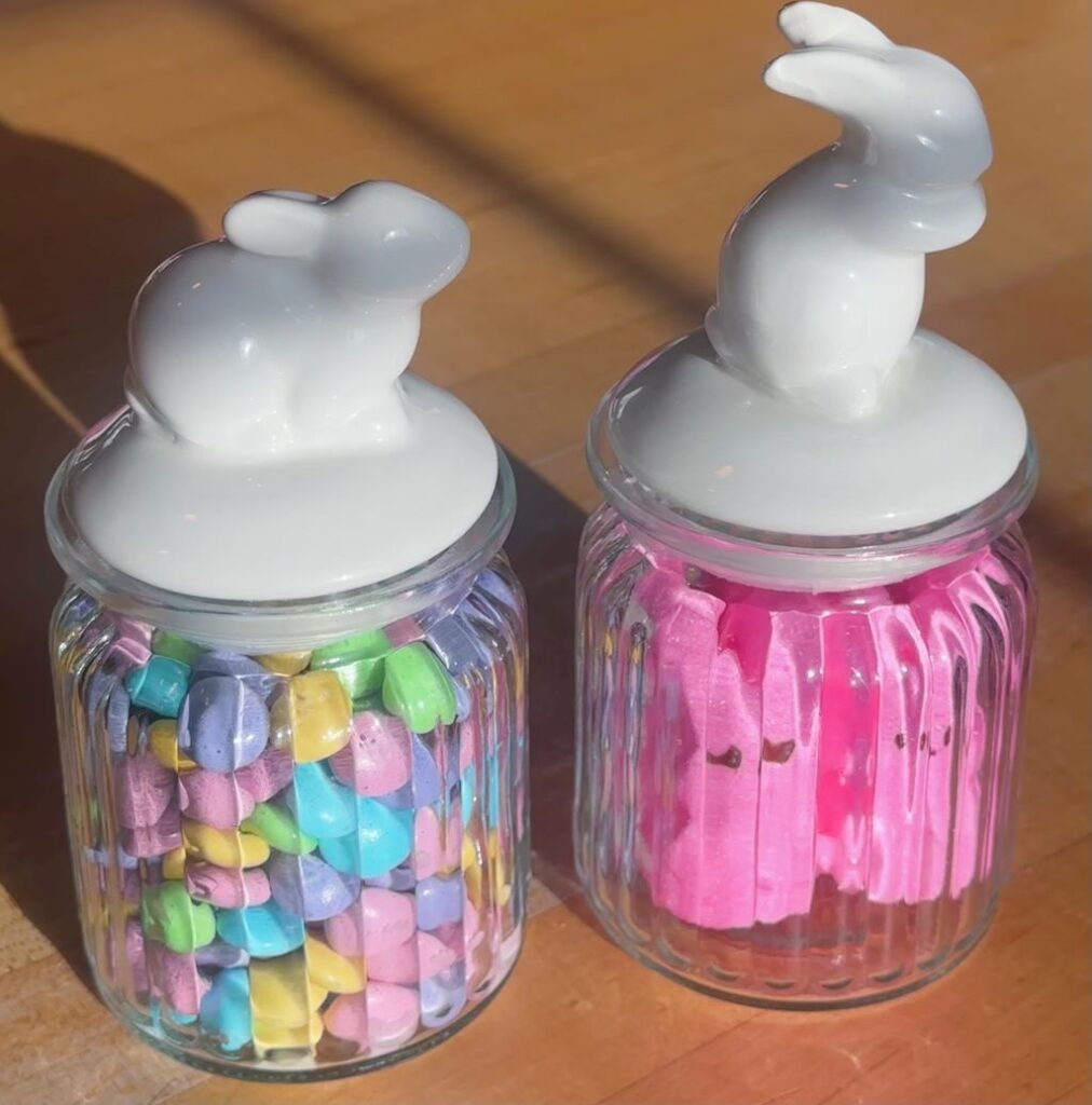 A picture of two glass jars with Easter bunny lids containing pastel M&Ms and pink bunny Peeps, sitting on a kitchen counter.