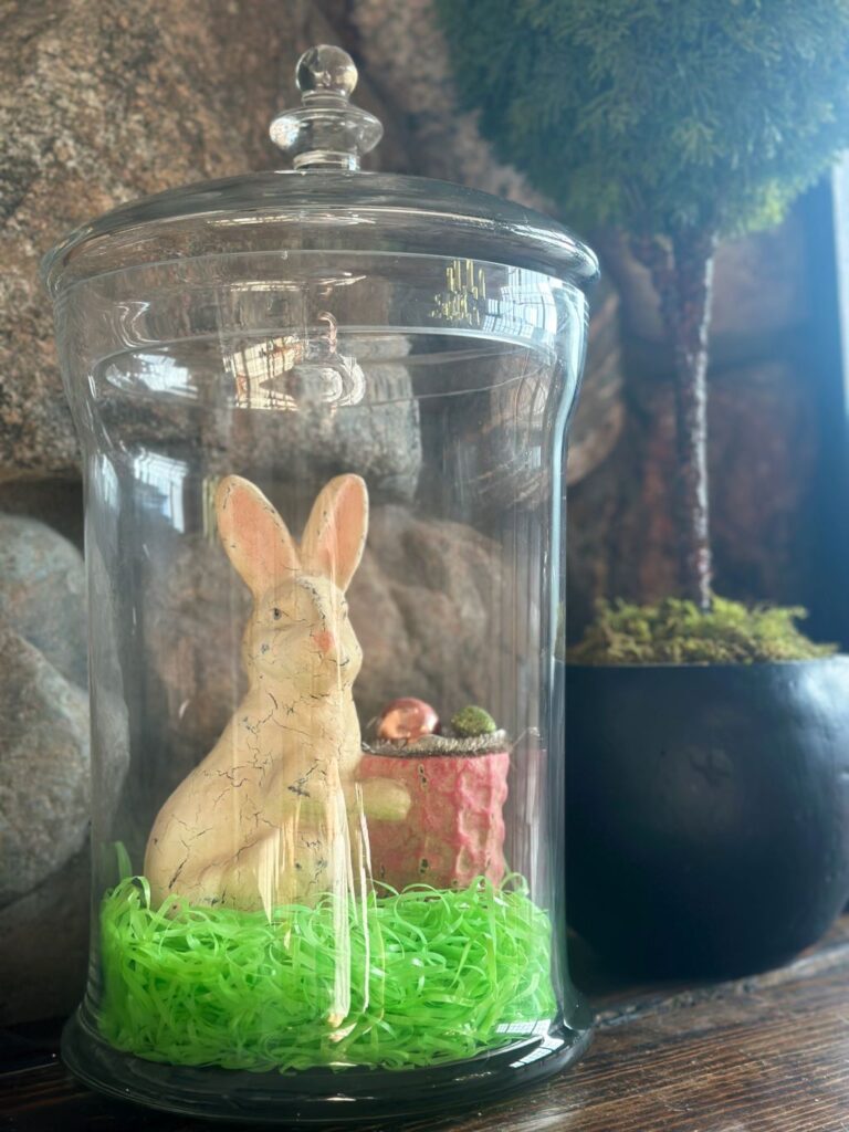A figurine of a white bunny with a little basket inside of a large apothecary jar with a fake boxwood seen in the background.