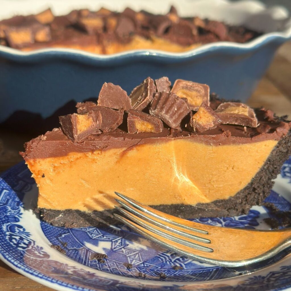 A close up of a slice of no-bake peanut butter pie on a small dessert plate with a fork, showcasing the Oreo crust, peanut butter filling, and ganache layers with toppings.
