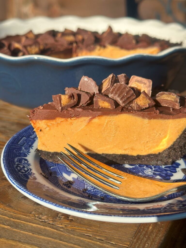 A close up of a slice of no-bake peanut butter pie on a small dessert plate with a fork, showcasing the Oreo crust, peanut butter filling, and ganache layers with toppings.