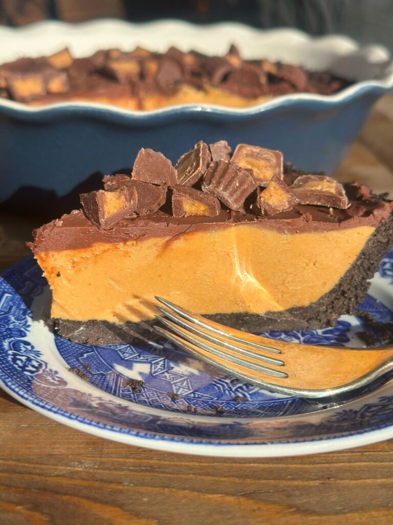 A close up of a slice of no-bake peanut butter pie on a small dessert plate with a fork, showcasing the Oreo crust, peanut butter filling, and ganache layers with toppings.