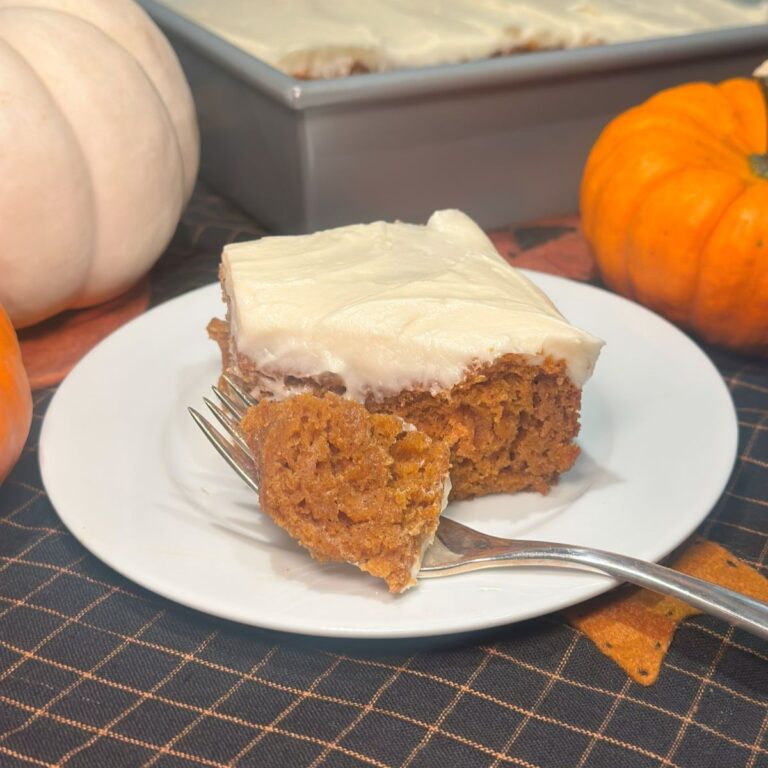 A slice of pumpkin snack cake on a small plate with a bite sitting on a fork, surrounded by small pumpkins.